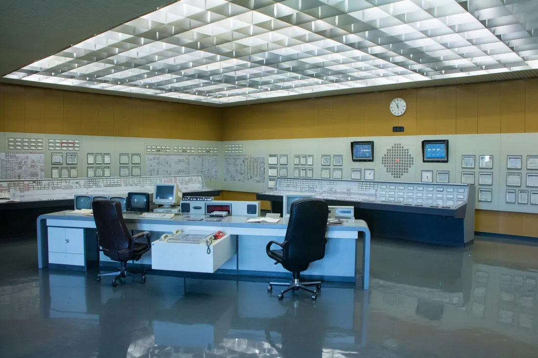Une salle de contrôle dont les murs sont couverts de tableaux d’instruments, une horloge posée parmi eux, les bureaux et les sièges laissés vides sous un plafond lumineux.