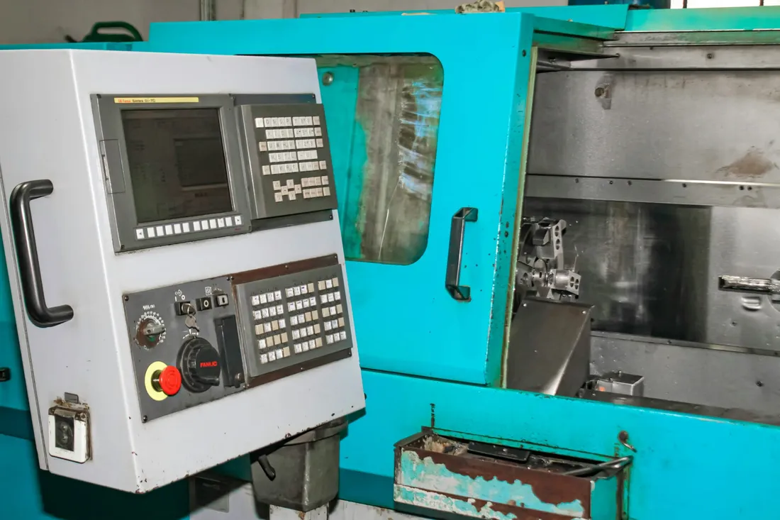 Un pupitre de commande fixé au flanc d’une machine-outil dans un atelier, son écran et son clavier au-dessus d’un arrêt d’urgence rouge, le carter de la machine et sa vitre derrière.