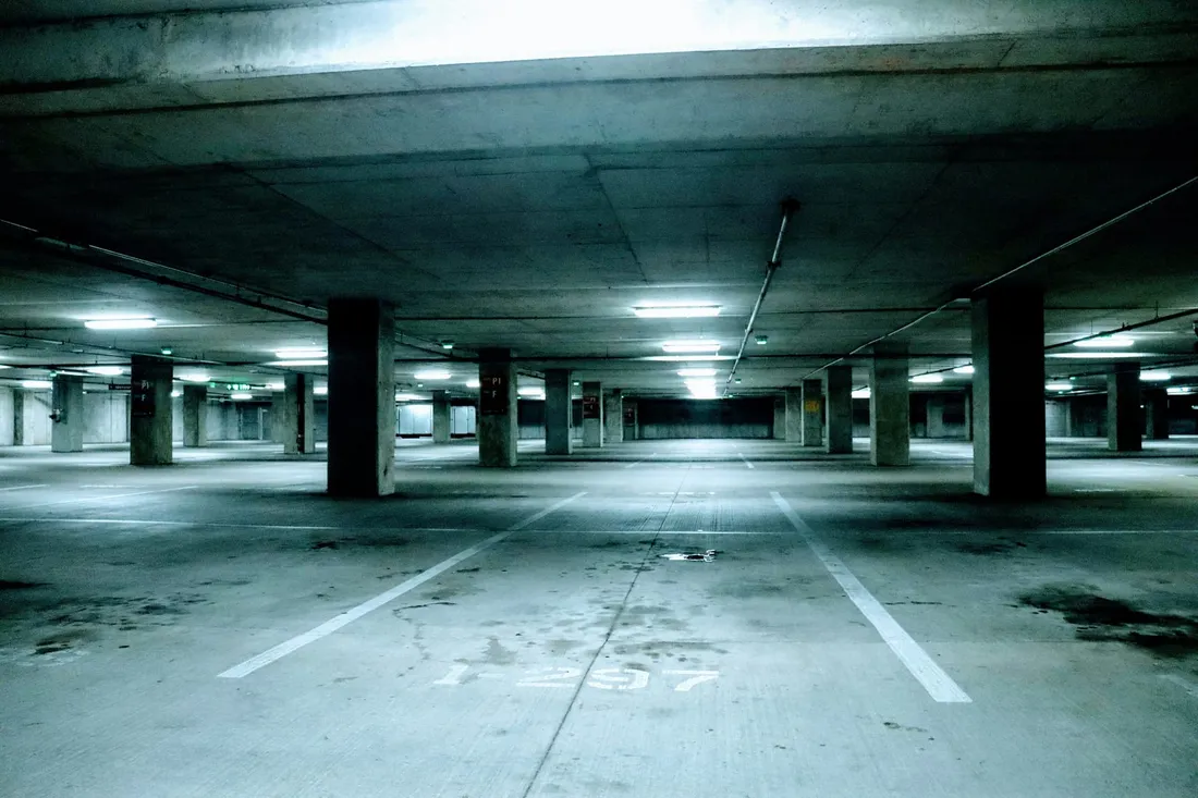 Un parking couvert dont les places sont tracées au sol et vides, les lignes blanches s’éloignant entre les piliers sous une rampe de plafonniers, un numéro peint au pochoir sur le béton au premier plan.